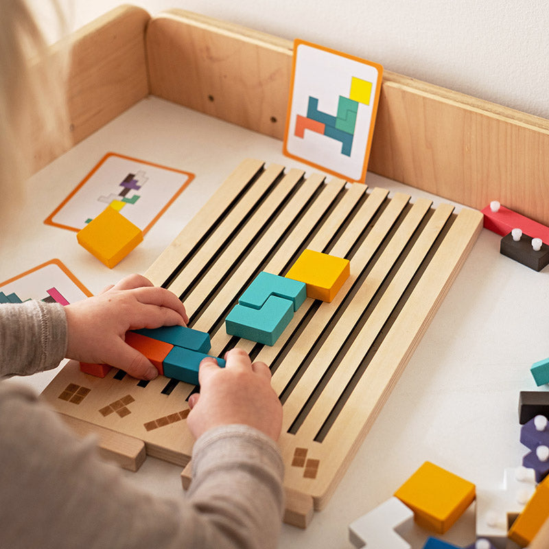 Kind legt met beide handen gekleurde blokken in de groeven van het houten bord, met opdrachtkaarten ernaast. 3