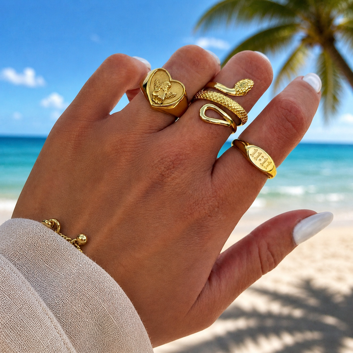 Hand op het strand met de gouden hartring met engeltje, samen gedragen met een slangenring en een ovale ring. 3