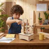 Kind in schort bouwt een houten handpomp met spuit, slangetjes en waterbakje, omringd door gereedschap en hout. 3