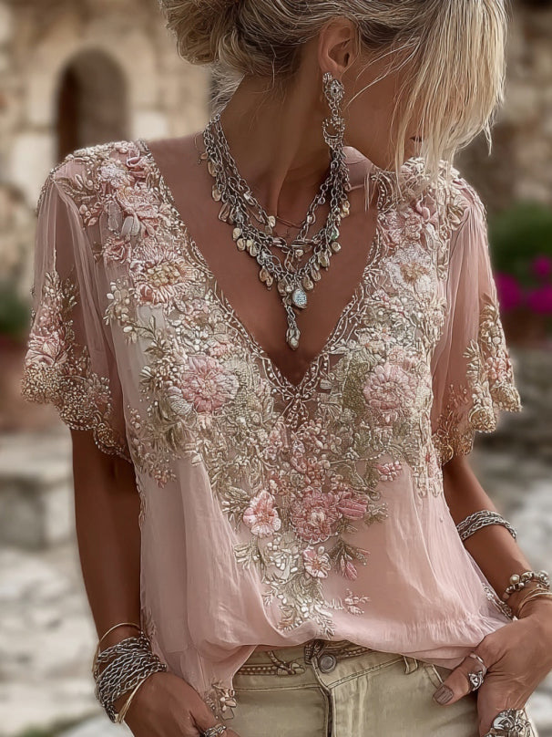 Vrouw draagt roze blouse met bloemenborduursel rond de V-hals en op de korte mouwen, gecombineerd met zilverkleurige kettingen en armbanden. 1