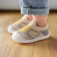 Zijaanzicht van beige gebreide babyschoentjes met gele streepdetails en grijs mesh, gedragen op een houten vloer. 9