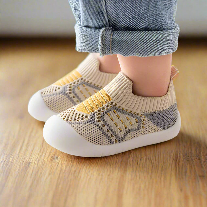 Zijaanzicht van beige gebreide babyschoentjes met gele streepdetails en grijs mesh, gedragen op een houten vloer. 9
