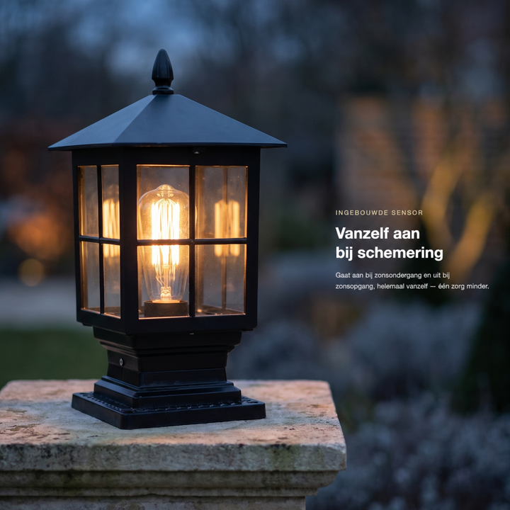 Brandende zwarte pilaarlamp op een stenen pilaar in de schemering, met een tuin vaag op de achtergrond. 2