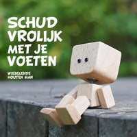 Houten figuurtje met zwarte oogjes zit met bungelende benen op een stenen rand, met groene bladeren wazig op de achtergrond. 15
