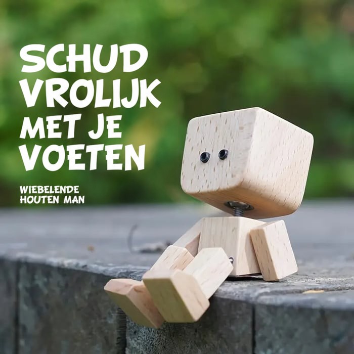 Houten figuurtje met zwarte oogjes zit met bungelende benen op een stenen rand, met groene bladeren wazig op de achtergrond. 15