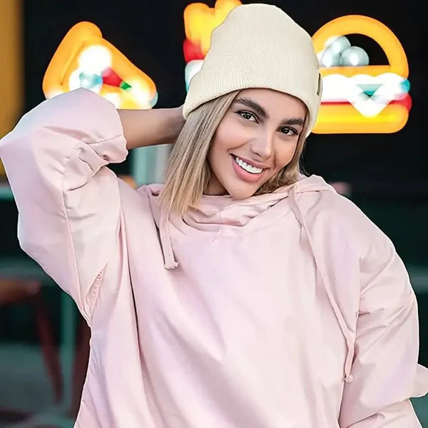Lachende vrouw met roze hoodie draagt een crèmekleurige muts tegen een achtergrond met neonlichten 2