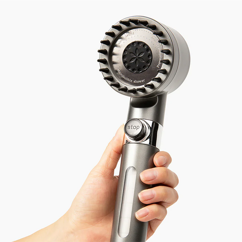 Hand houdt de grijze handdouche vast en toont de voorkant met zwarte siliconen noppen en de stopknop op het handvat. 3