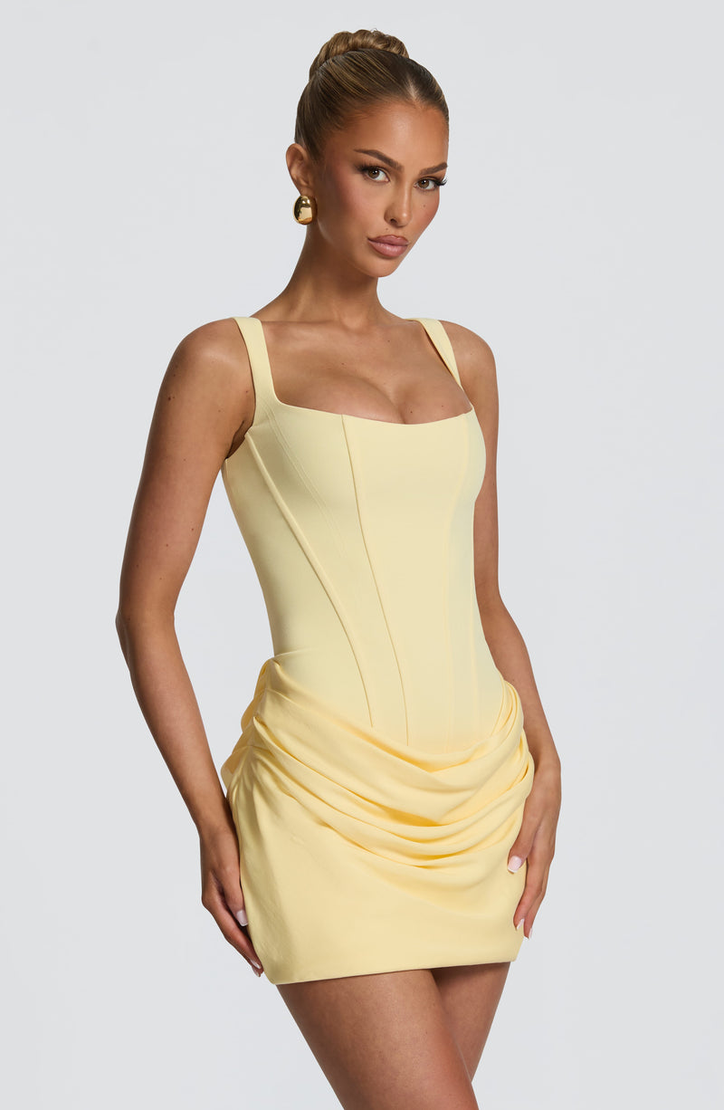 Lichtgele mini-jurk in driekwartaanzicht op een model, met gedrapeerde plooien over de heup en corsetnaden in het lijfje. 24