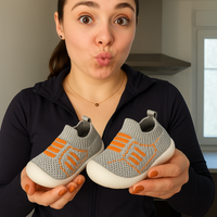 Vrouw houdt een paar grijze gebreide babyschoentjes met oranje streepdetails en witte zool omhoog naar de camera. 2