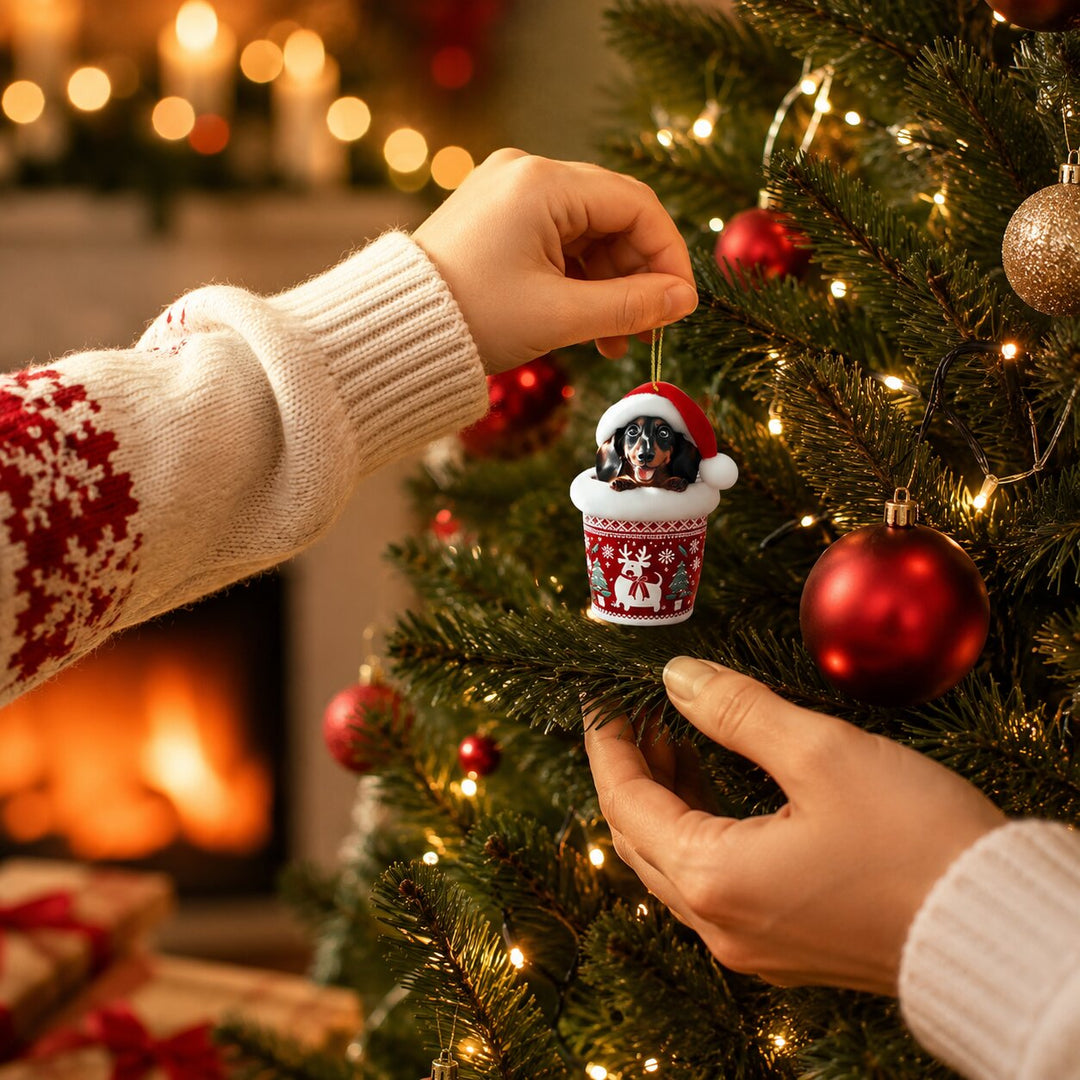 Close-up van een hand die een teckelhanger in een rood-wit kopje met rendiermotief aan een kerstboomtak hangt. 4