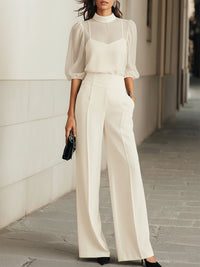 Model in crème set met doorschijnend bovenstuk, hemdje en wijde pantalon met hoge taille, staand langs een muur buiten. 2