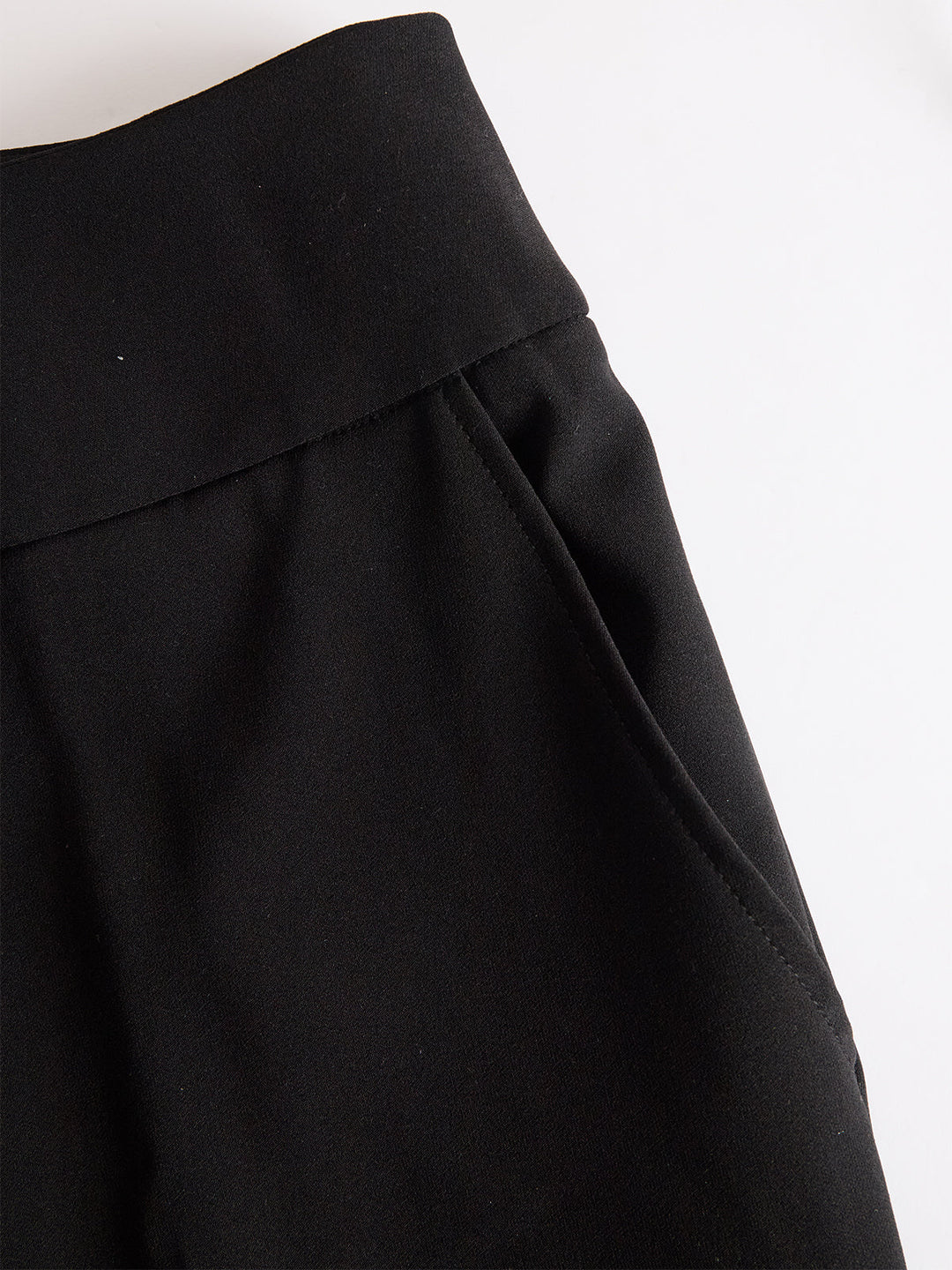 Detail van de zwarte broek met brede tailleband en schuin ingestoken zak, plat neergelegd op een witte ondergrond. 5