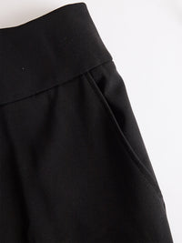 Detail van de zwarte broek met brede tailleband en schuin ingestoken zak, plat neergelegd op een witte ondergrond. 5