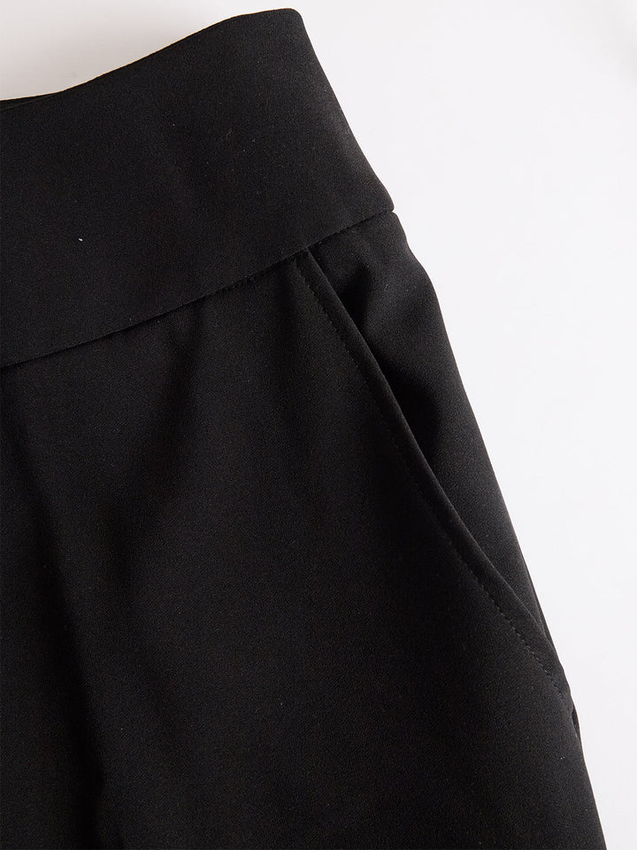 Detail van de zwarte broek met brede tailleband en schuin ingestoken zak, plat neergelegd op een witte ondergrond. 5