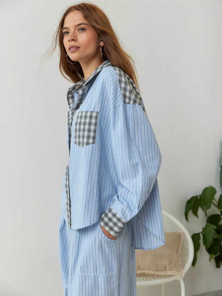 Zijaanzicht van model in blauw gestreept overhemd met grijs geruite details en bijpassende broek, naast een wit rotanstoeltje en plant. 3