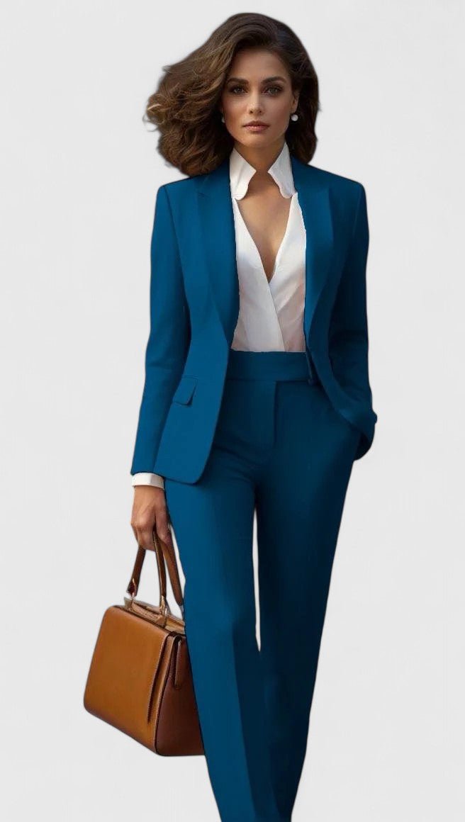 Model draagt het kostuum in petrolblauw, met witte blouse met golvende kraag en een bruine handtas. 2