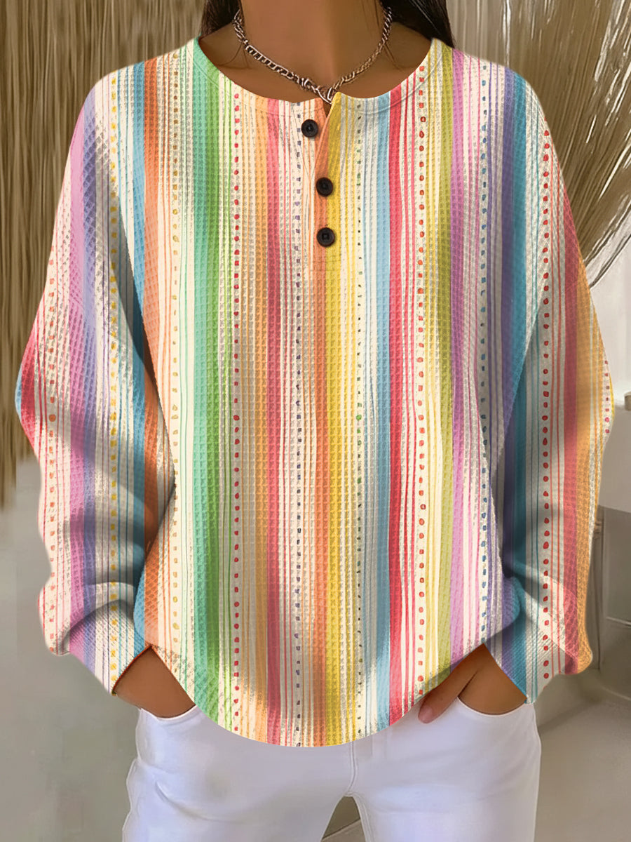 Wafeltrui met knoopjes en verticale regenboogstrepen met stippellijnen in vrolijke kleuren, gedragen met witte broek. 10