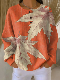Oranje wafeltrui met knoopjes en grote beige esdoornbladeren als print, gedragen met witte broek. 14