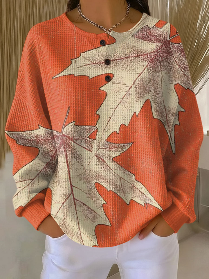 Oranje wafeltrui met knoopjes en grote beige esdoornbladeren als print, gedragen met witte broek. 14