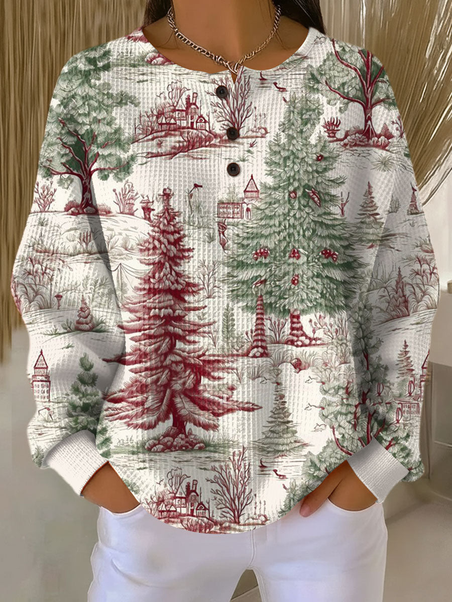Crèmewitte wafeltrui met knoopjes en een rood-groene winterprint van dennenbomen, huisjes en kale bomen, gedragen met witte broek. 22