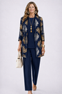 Model in donkerblauwe set met gouden bladprint op het open vest, effen top en broek, beige tas in de hand. 7
