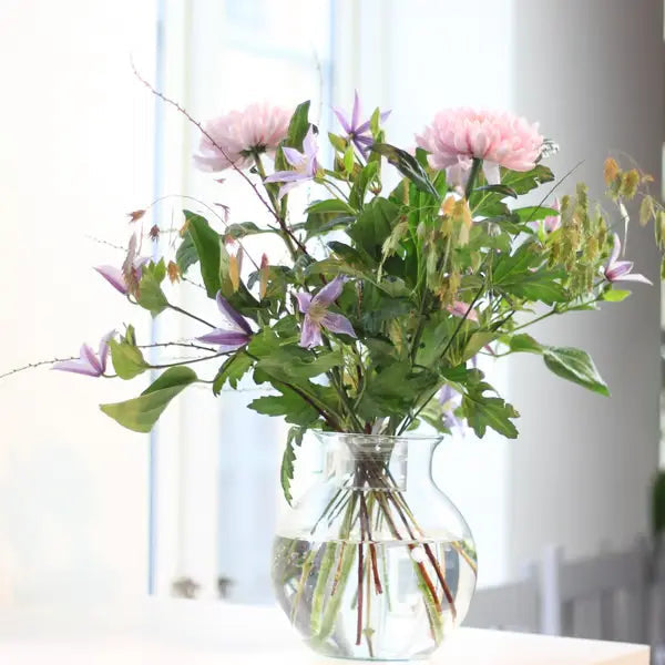 Boeket met roze bloemen en paarse clematis staat luchtig en rechtop in een heldere glazen vaas met water bij een raam. 4
