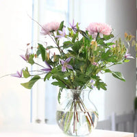 Boeket met roze bloemen en paarse clematis staat luchtig en rechtop in een heldere glazen vaas met water bij een raam. 4
