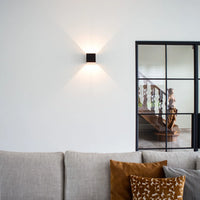 Zwarte wandlamp op een witte muur boven een grijze bank met okergele kussens, naast een stalen glazen deur. 10
