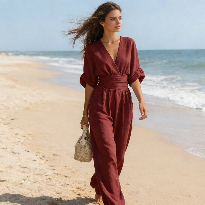 Vrouw in donkerrode jumpsuit wandelt op het strand met wapperend haar en een rieten tas in haar hand. 7