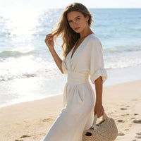 Vrouw in crèmewitte jumpsuit staat op het strand met een rieten tas, close-up van de V-hals en brede taille. 12