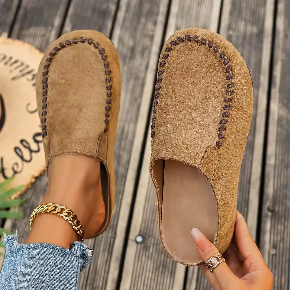 Bovenaanzicht van instapklompen in camel suèdelook met bruine gevlochten stiksels, de ene aan de voet, de andere in de hand met zicht op de binnenzool. 1