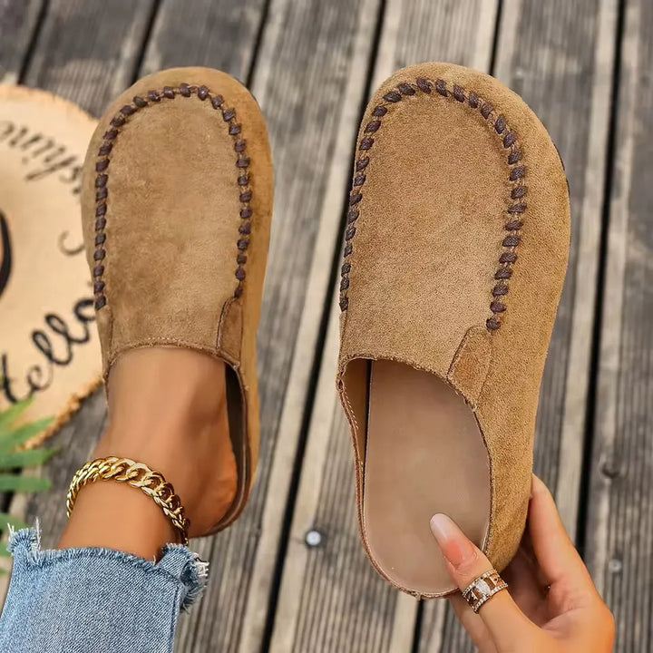 Bovenaanzicht van instapklompen in camel suèdelook met bruine gevlochten stiksels, de ene aan de voet, de andere in de hand met zicht op de binnenzool. 1