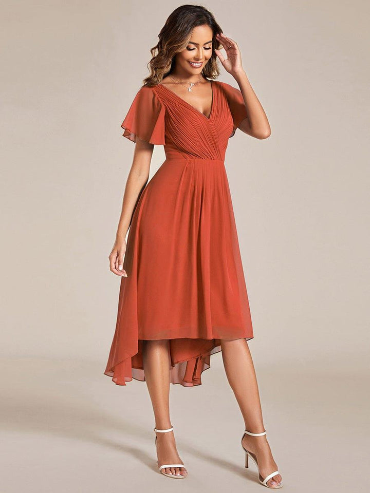 Terracotta jurk in driekwartaanzicht, model raakt haar haar aan terwijl de chiffon rok zacht meebeweegt. 96
