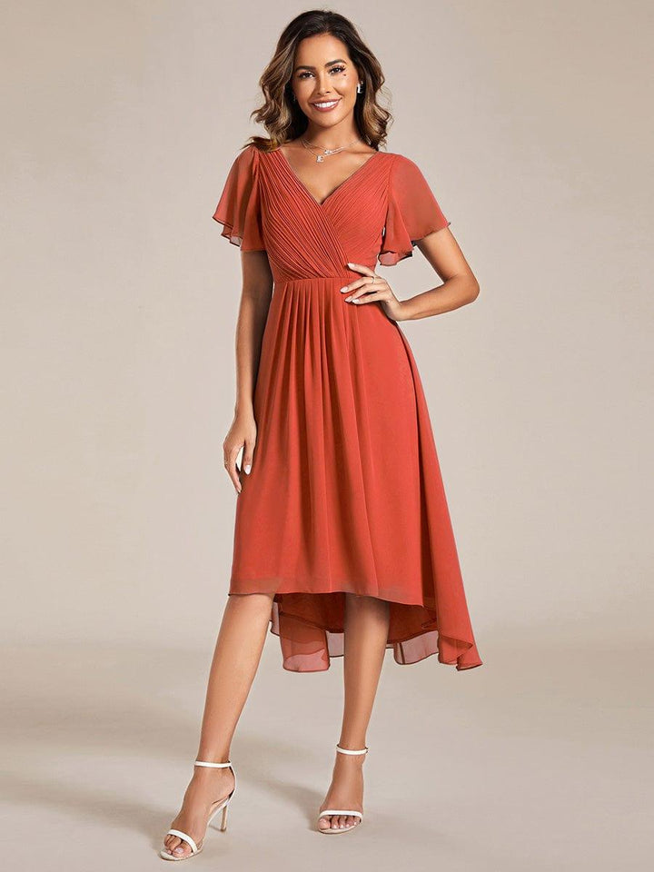 Model in terracotta-oranje chiffon jurk met geplooide V-hals en korte fladdermouwen, hand op de heup, witte sandalen. 92