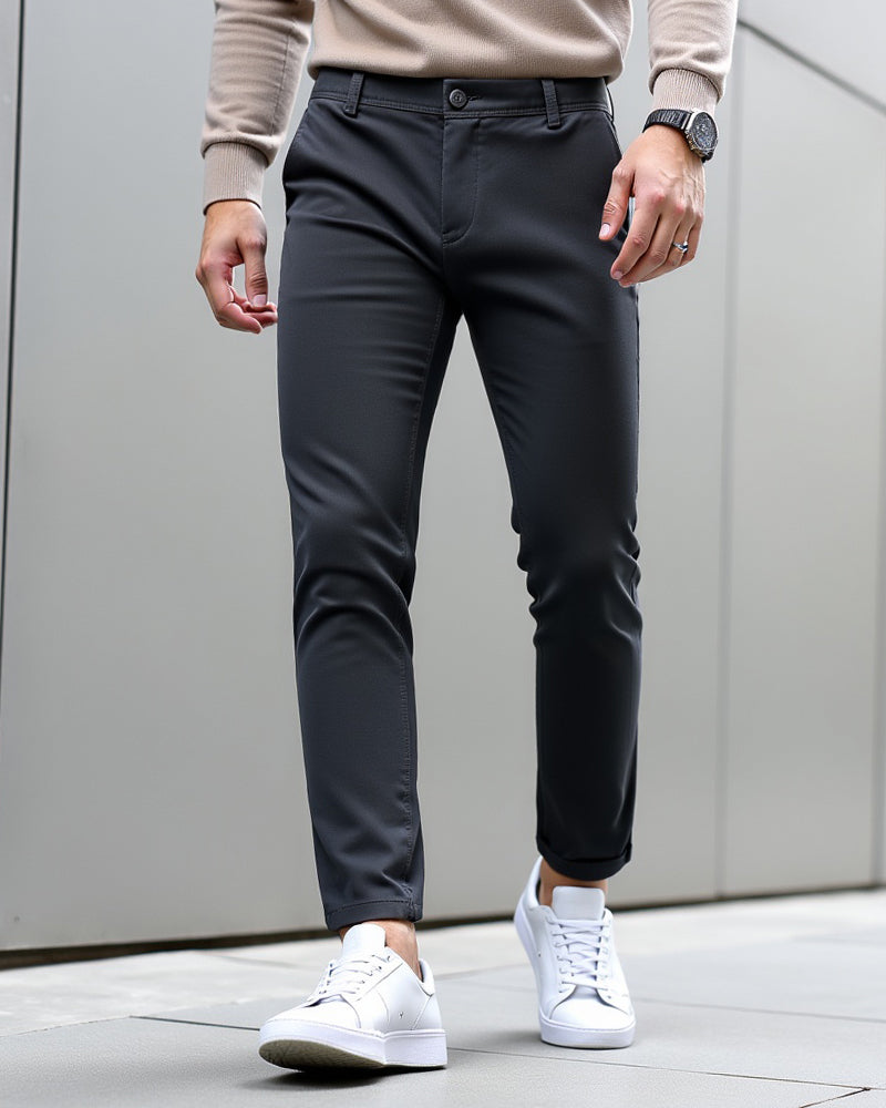Donkergrijze chino met omgeslagen pijpen gedragen met beige trui, horloge en witte sneakers voor een grijze wand. 9
