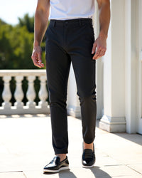 Donkergrijze chino gedragen op een zonnig terras met wit shirt en zwarte loafers, tegen een witte balustrade. 7