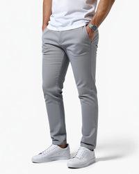 Lichtgrijze slim fit chino van opzij gedragen door een model met witte sneakers, wit shirt en horloge, hand in zak. 1