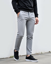 Lichtgrijze chino gedragen met zwarte sweater en zwarte sneakers, model leunt tegen een witte muur buiten. 2