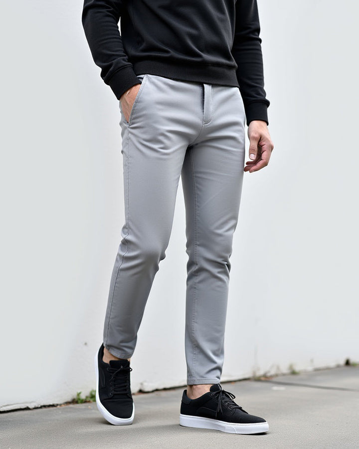 Lichtgrijze chino gedragen met zwarte sweater en zwarte sneakers, model leunt tegen een witte muur buiten. 2