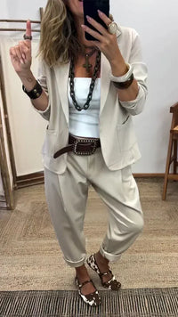 Vrouw in beige kostuum met blazer met opgestikte zakken en ruime broek, gedragen met wit topje, grote halsketting en bruine riem. 2