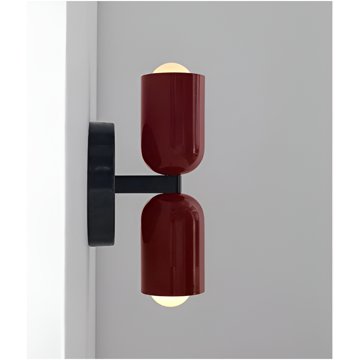 Zijaanzicht van een wandlamp met twee rode lampkoppen op een zwarte arm en ronde zwarte muurplaat, de peertjes lichten warm op. 17
