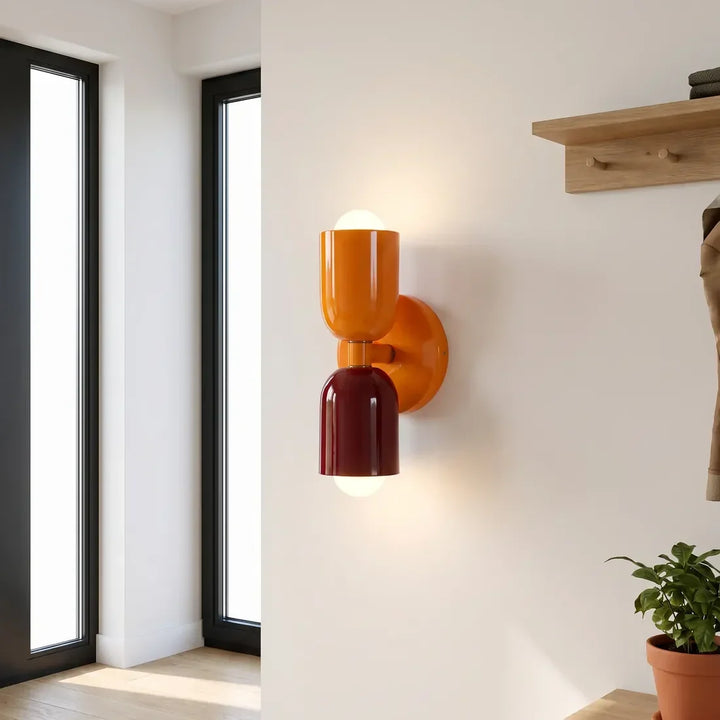 Wandlamp in oranje en bordeauxrood naast hoge ramen in een hal, met een houten kapstokplank en plant. 11
