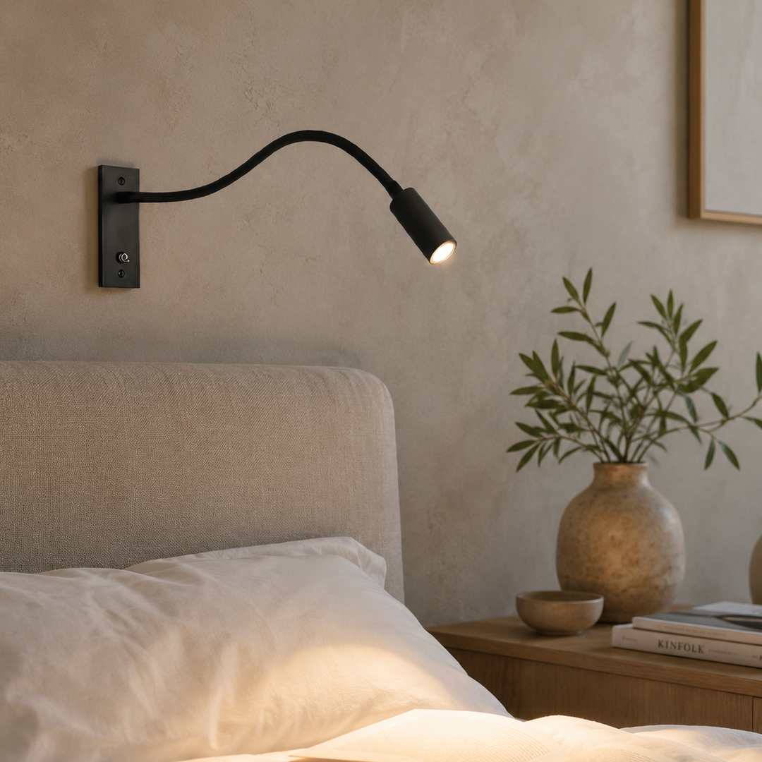 Zwarte wandlamp met brandend licht boven een bed met linnen kussens, naast een nachtkastje met vaas en boeken. 3