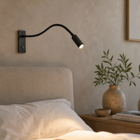 Zwarte wandlamp met brandend licht boven een bed met linnen kussens, naast een nachtkastje met vaas en boeken. 3