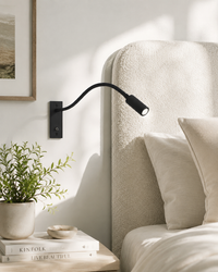 Zwarte leeslamp aan de muur naast een bed met crème bouclé hoofdbord, kussens en een plant op het nachtkastje. 2