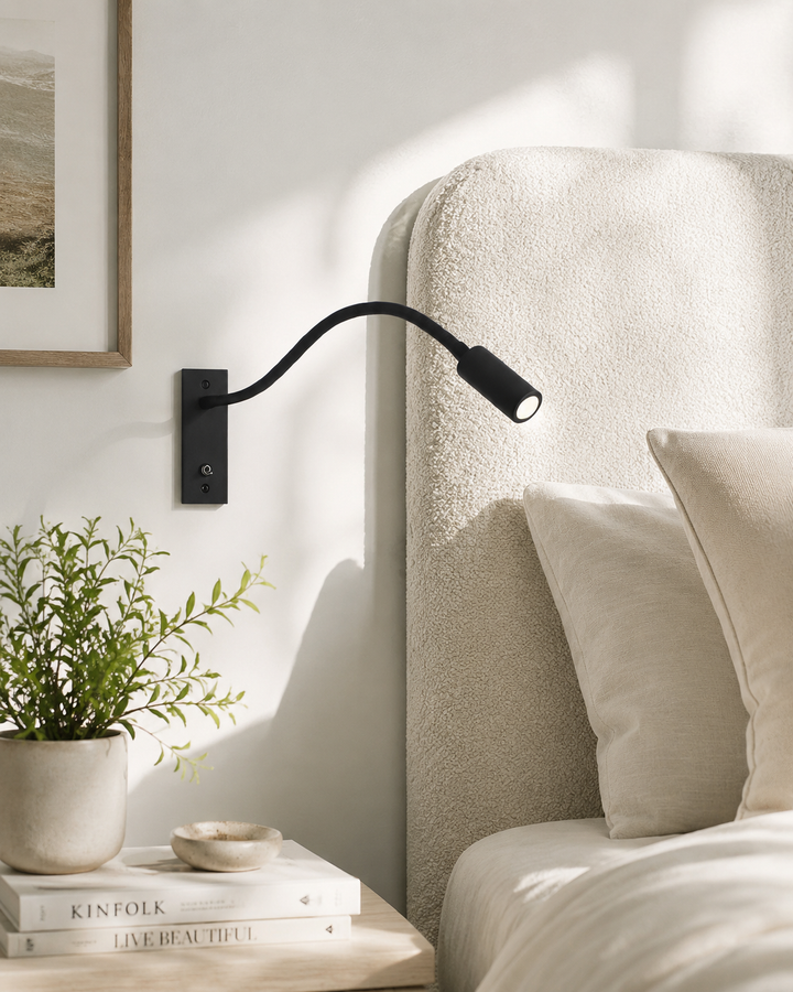 Zwarte leeslamp aan de muur naast een bed met crème bouclé hoofdbord, kussens en een plant op het nachtkastje. 2