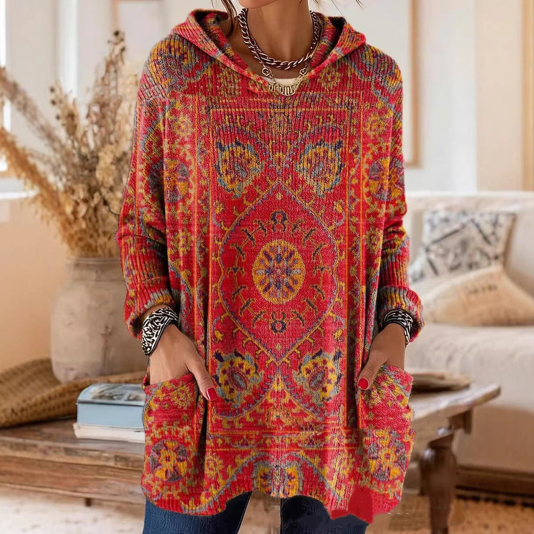 Model draagt de rode tuniek met capuchon en tapijtachtige print, handen in de zakken, in een woonkamer met gedroogde bloemen. 1