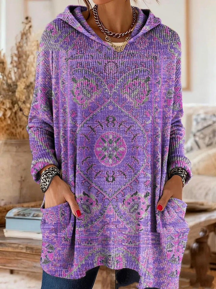 Dezelfde tuniek met capuchon in paars met roze en groene tapijtprint, gedragen met handen in de voorzakken. 2