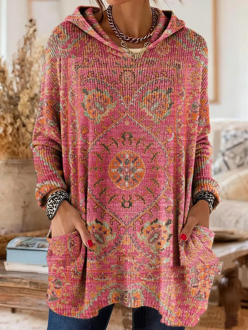Roze tuniek met capuchon en oranje en groene tapijtprint, gedragen door een model met handen in de voorzakken. 6