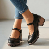 Zwarte Mary Jane-schoenen met brogue-perforatie en bandje aan de voeten van een model in spijkerbroek, close-up op effen ondergrond 1